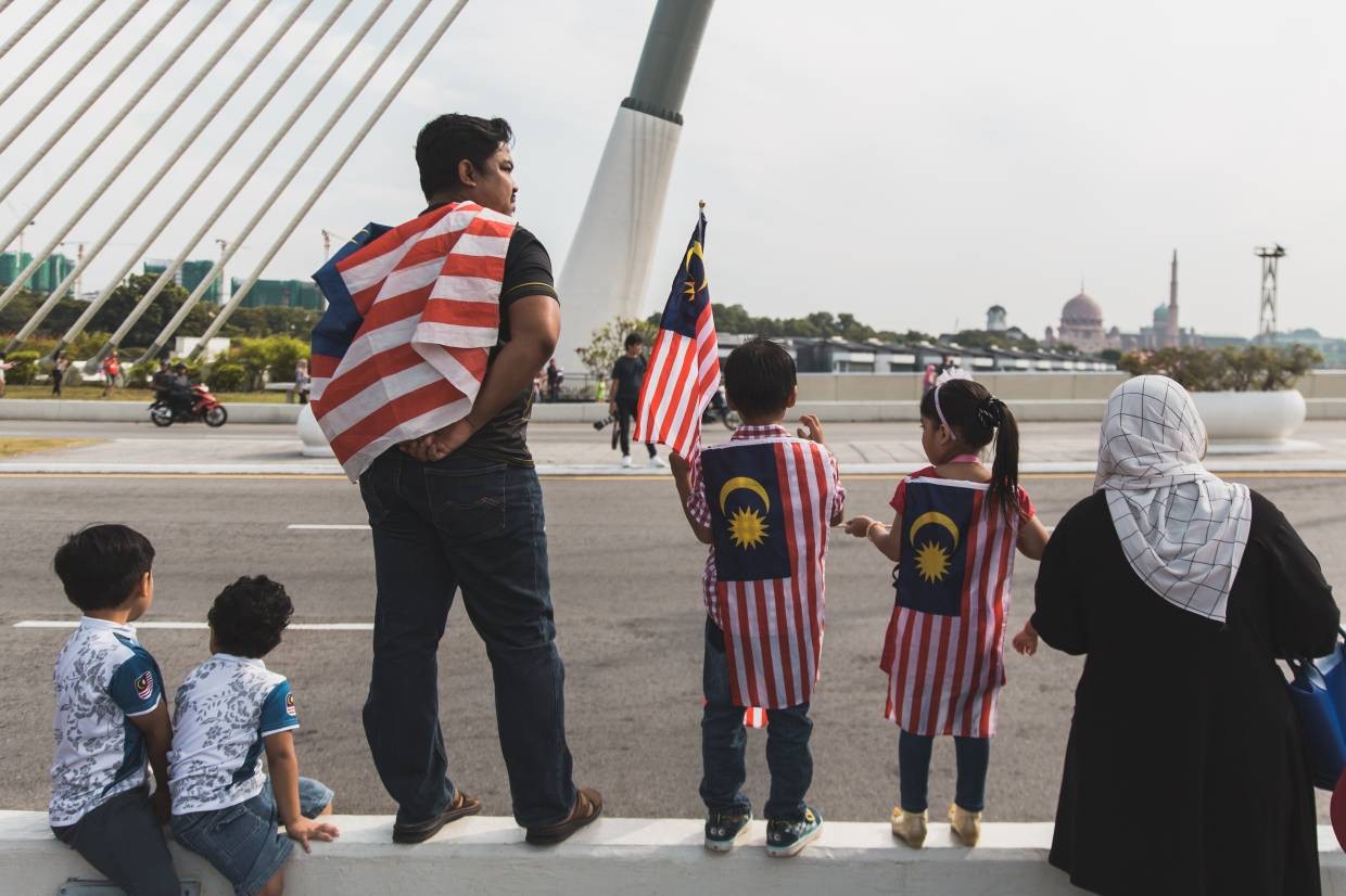 A father and his two small children – all wearing the Jalur Gemilang flag – wait for the parade to pass by.