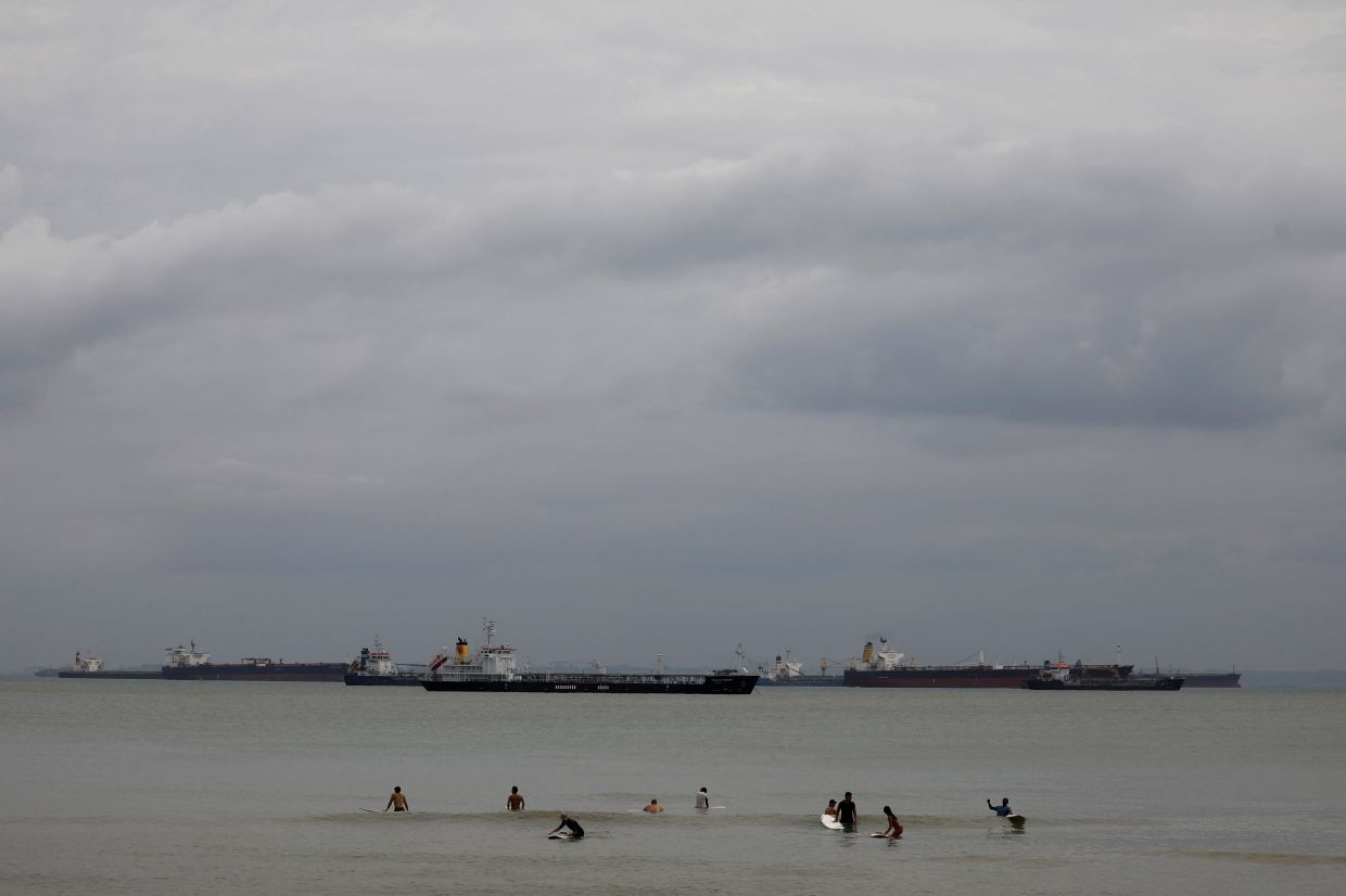 Surfers wait to catch a wave along a beach surrounded by tankers in Singapore. - Reuters