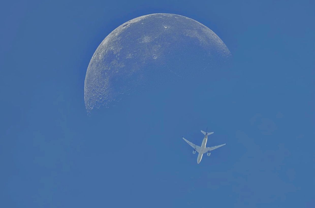 Photographing the moon during the blue hour with a plane flying near the moon in the foreground adds depth to the photo.- Pic by Eddie Chua