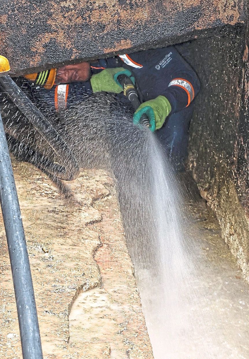 An Alam Flora worker squeezing into a tight, partially covered drain to clear hardened grease and debris using a high-pressured water jet.