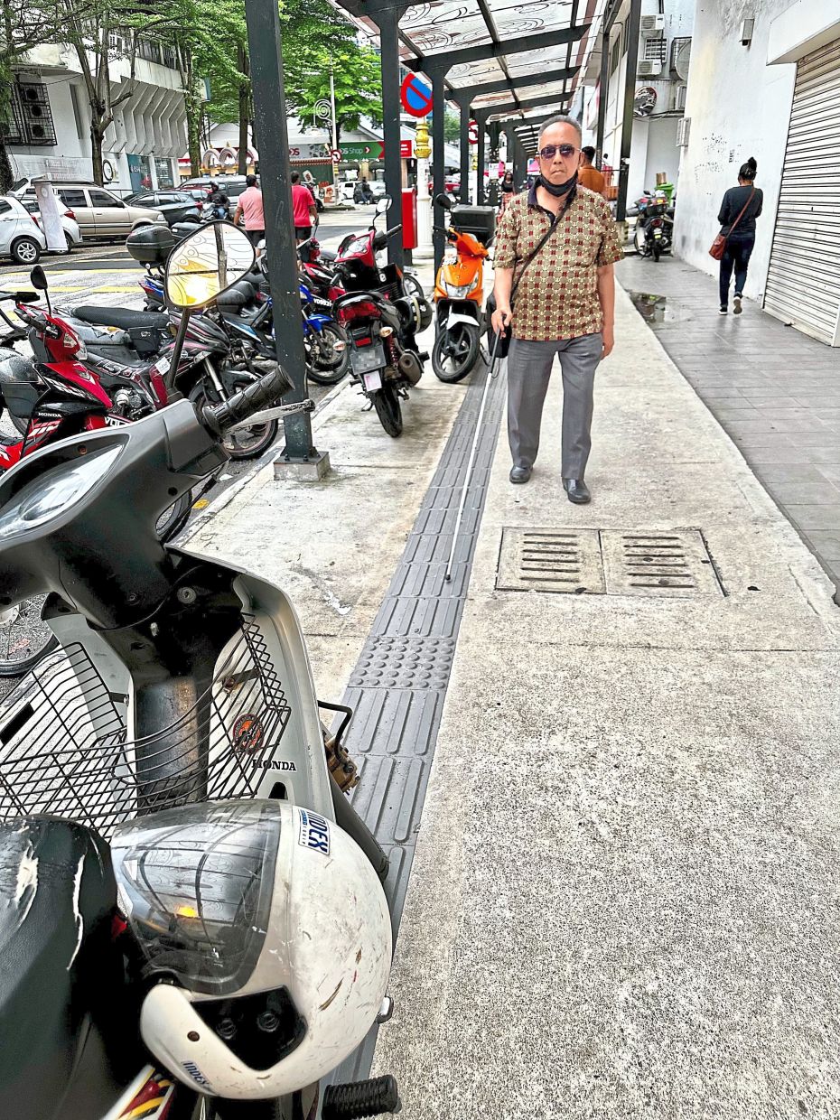 A visually impaired man navigates using the tactile path in Brickfields, which is obstructed by a parked motorcycle.