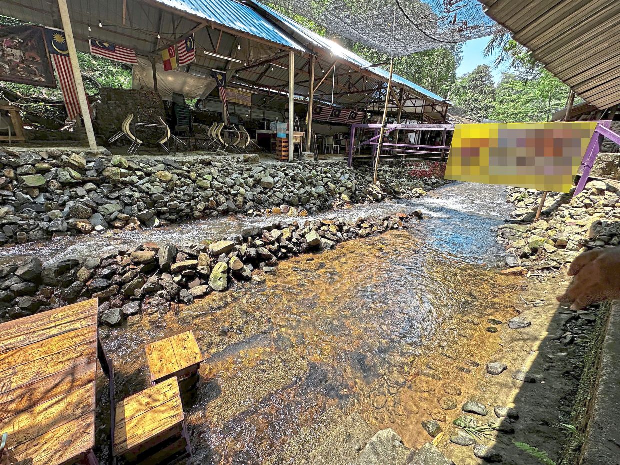 Wooden dining tables and stools placed in shallow Sungai Kemensah waters. Nearby, a bright yellow banner hung across the stream provides information on another business there.