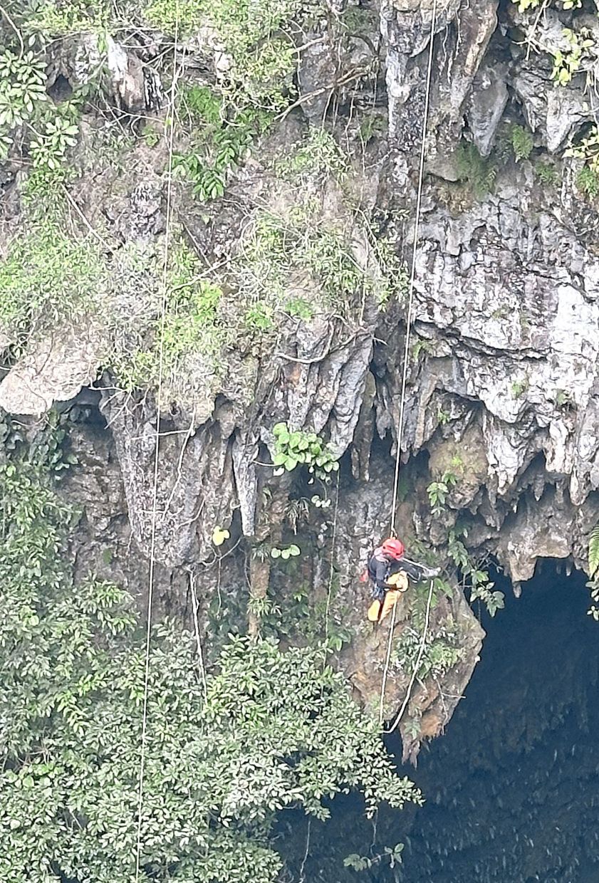 A cave climber lowering himself into the 60m-deep sinkhole, suspended on a rope against a backdrop of ancient rock and forest.