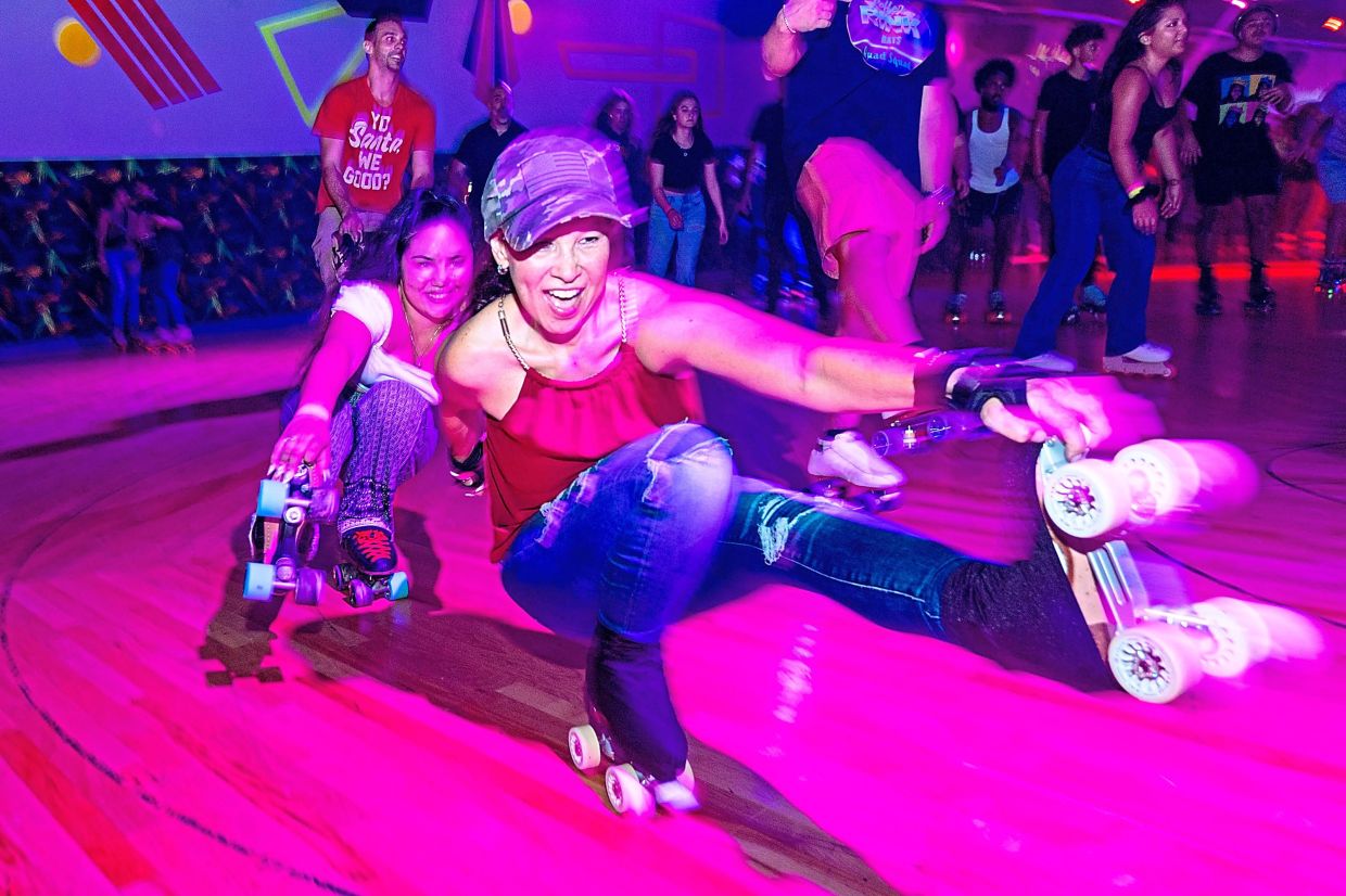 Two roller-skaters perform skate tricks around the roller rink.