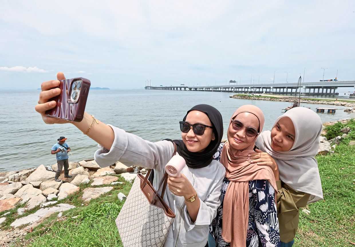 Halimah Munthe, 23, using a portable fan to keep cool while taking a wefie with her mother and sister, with Penang Bridge in the background.