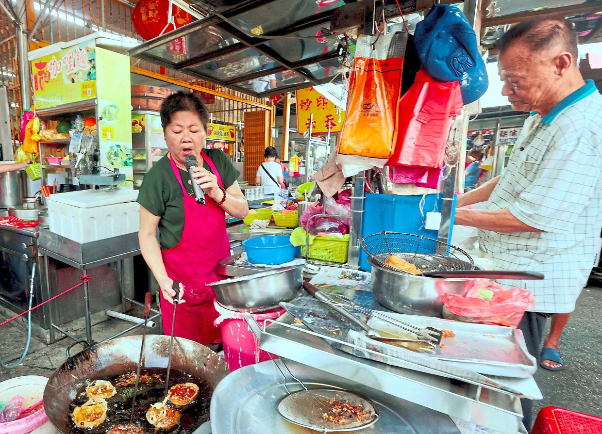 Tunes and treats: Lye belting out a song at her stall while frying snacks as the operator deals with customers in the Jelutong Market, Penang. — ZHAFARAN NASIB/The Star
