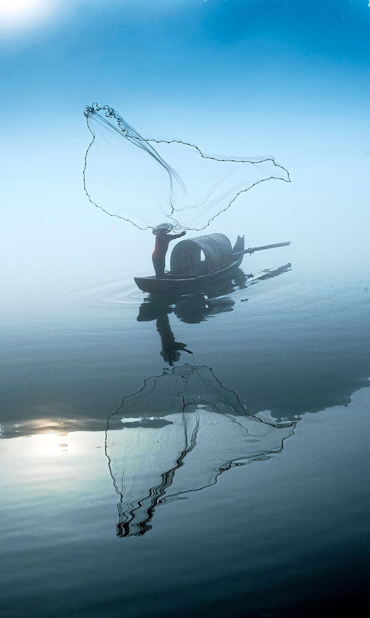 A fisherman casting his net over glassy waters in Chenzhou, China, his reflection rippling beneath the morning haze.