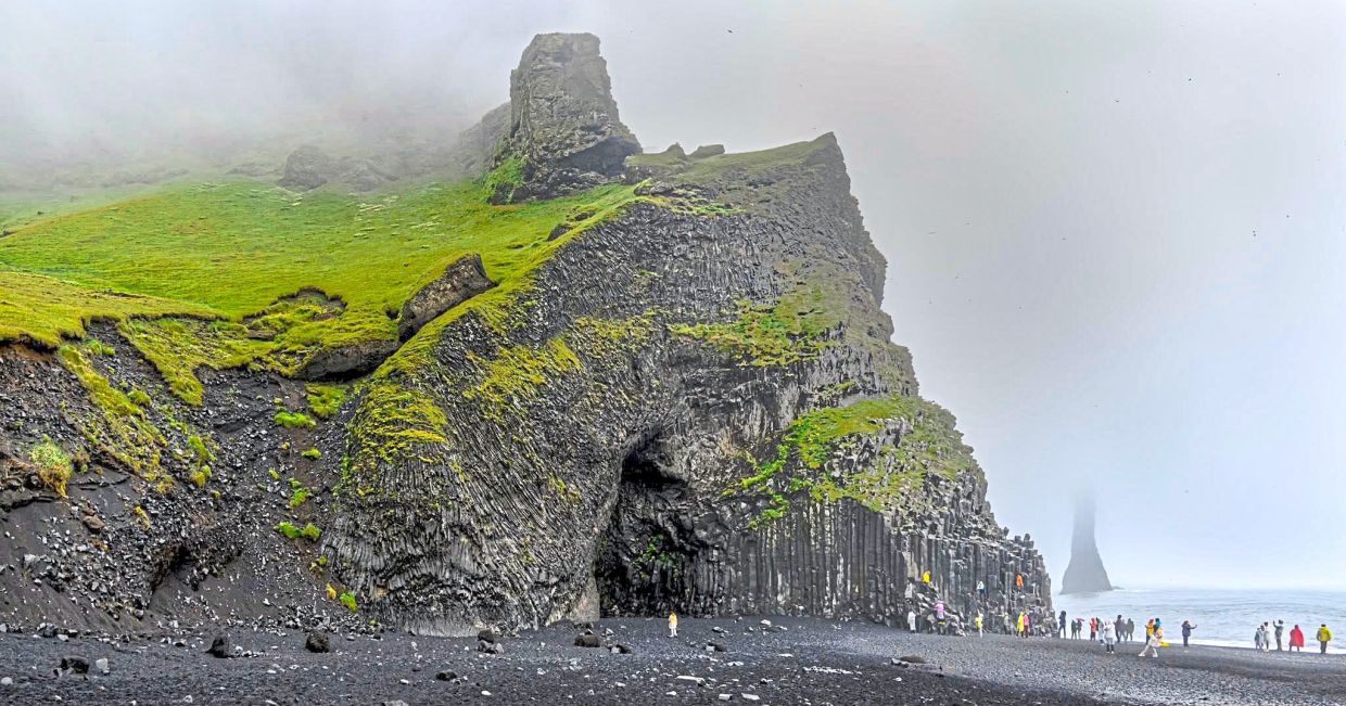 Reynisfjara’s black sand and basalt cliffs draw visitors year-round, where mist drifts around the Reynisdrangar sea stacks offshore.