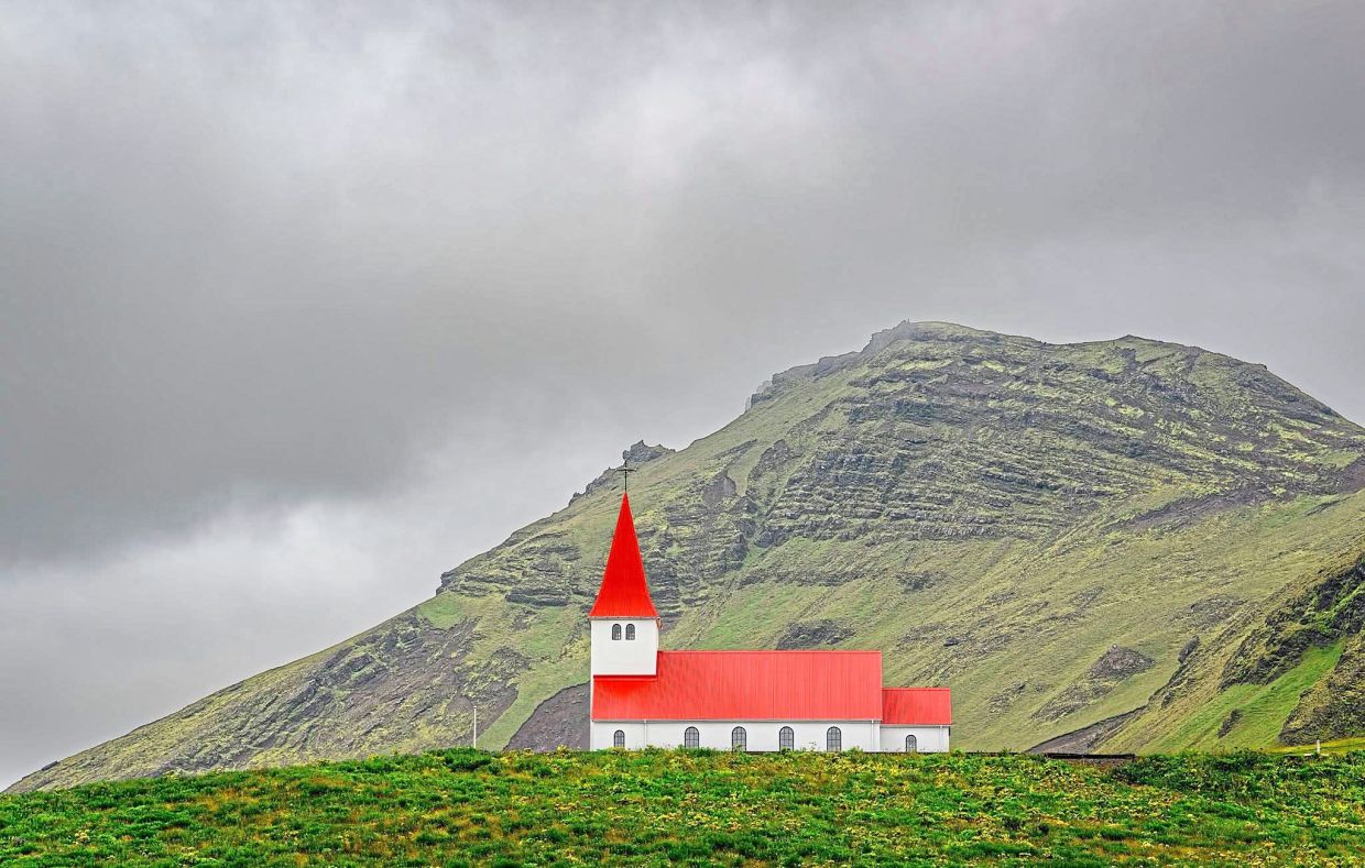 A solitary red-roofed church stands stark against Iceland’s moody volcanic ridges, a familiar landmark on the South Coast.