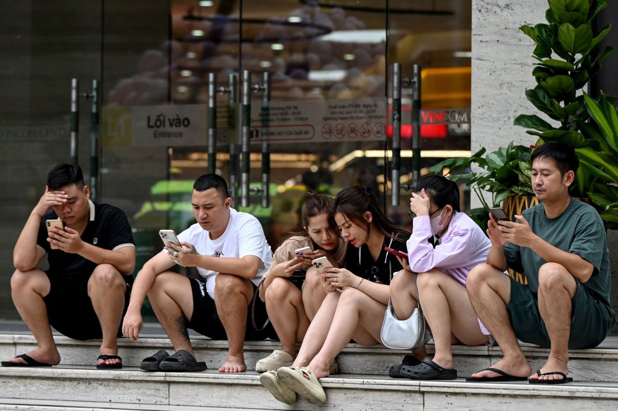 People use their phones as they relax at the entrance of a shopping mall in Hanoi on Monday, October 13, 2025. -- Photo by Nhac NGUYEN / AFP