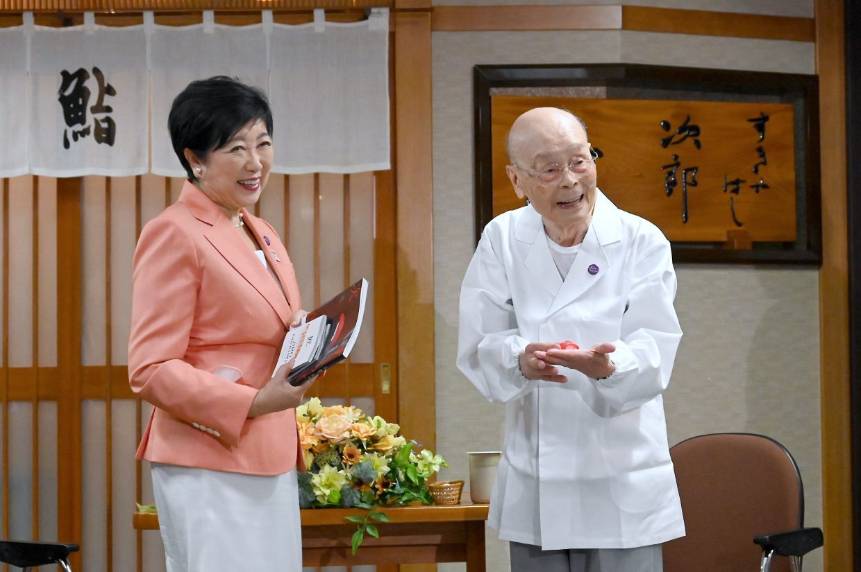 In this photo released by Bureau of Social Welfare, Tokyo Metropolitan Government, sushi legend Jiro Ono, right, shows a gift from Tokyo Gov. Yuriko Koike to celebrate his 100th birthday in front of his restaurant, Sukiyabashi Jiro in Tokyo on Sept. 18, 2025. (Bureau of Social Welfare, Tokyo Metropolitan Government via AP)