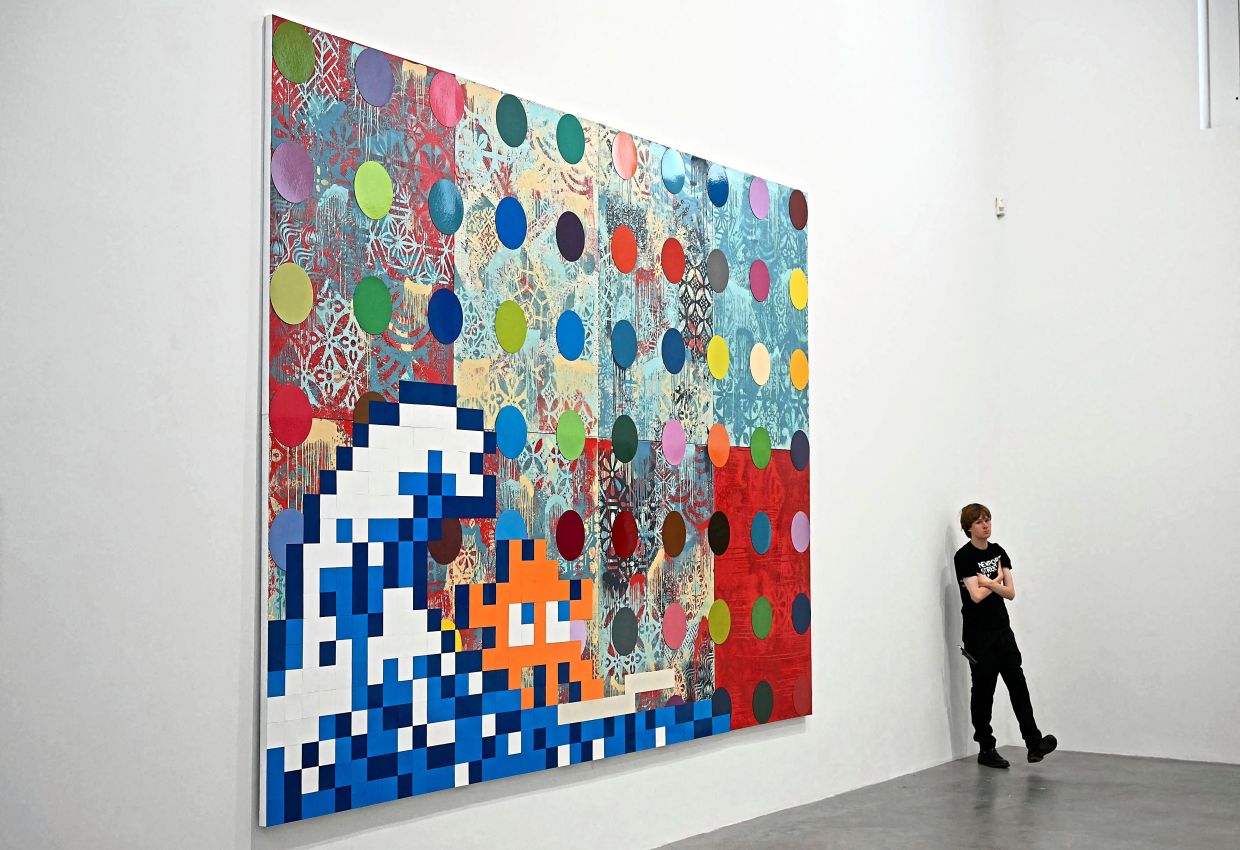 A gallery assistant stands next to the artwork Troubled Waters (2025) by Fairey, Hirst, and Invader during the Triple Trouble exhibition photo call in London.