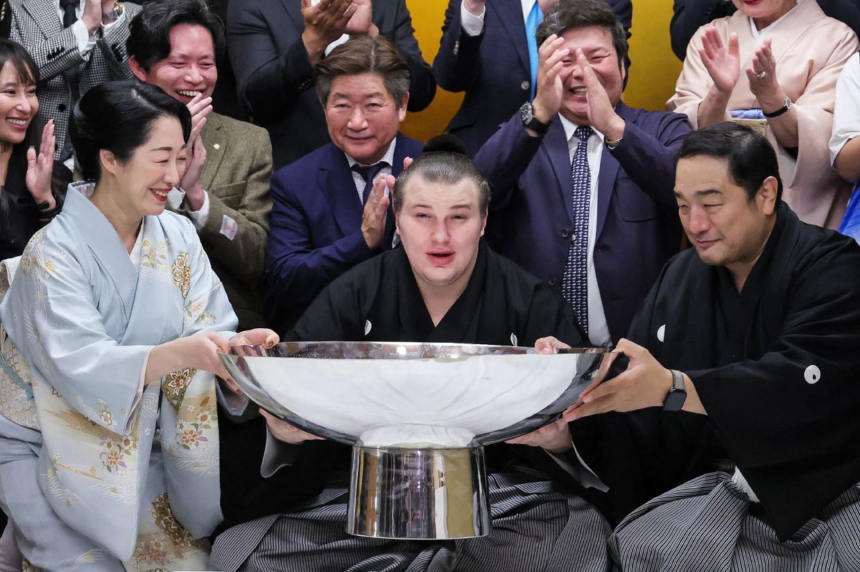 Ukrainian sumo wrestler Danylo Yavhusishyn (centre), also known by his Japanese ring name Aonishiki Arata, poses with a large cup to celebrate after winning the Grand Sumo Tournament in Kurume city of Fukuoka Prefecture on November 23, 2025. -- Photo by JIJI PRESS / AFP