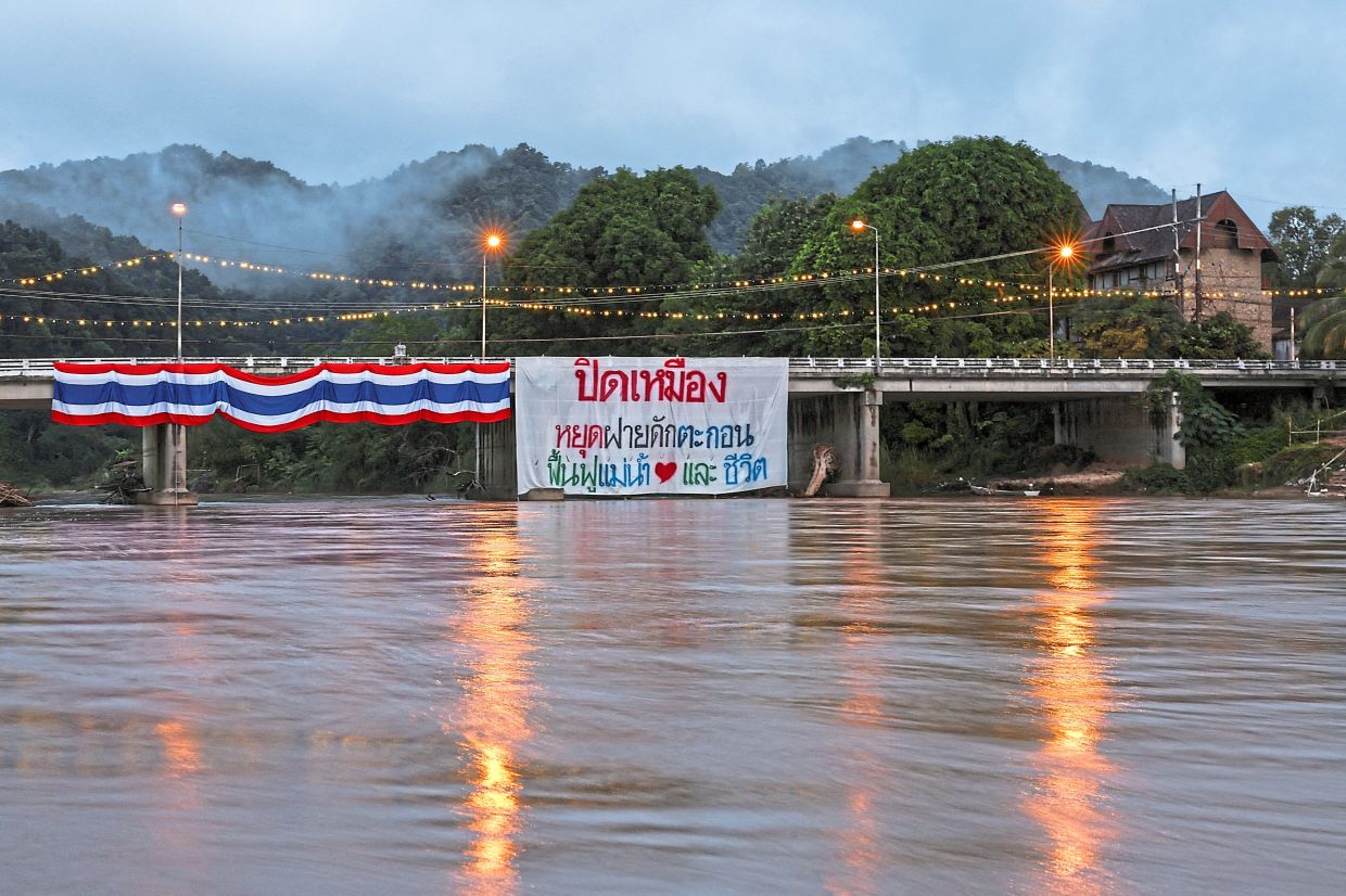 A sign reading 'Shut down the mines, stop the sediment trap, and restore the river and people's lives' hangs from a bridge over the Kok River.