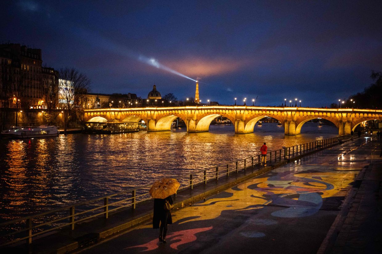 People walk along the banks of the Seine River with an illuminated Pont Neuf and the Eiffel Tower seen on the backgraund in Paris on November 29, 2025. — Photo: Dimitar DILKOFF / AFP