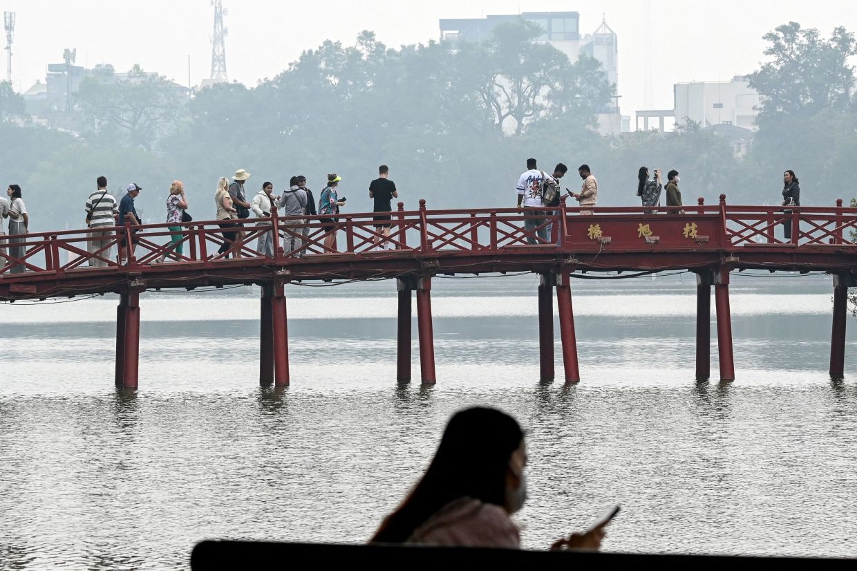 A girl wears a face mask as she uses her phone next to people crossing a bridge over West Lake amid heavy air pollution conditions in Hanoi on Dec 3, 2025. - AFP