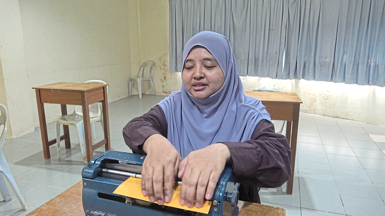 Rabiatul Adawiyah learning to read Braille using a manual Braille writer at the Taman Cahaya classroom.