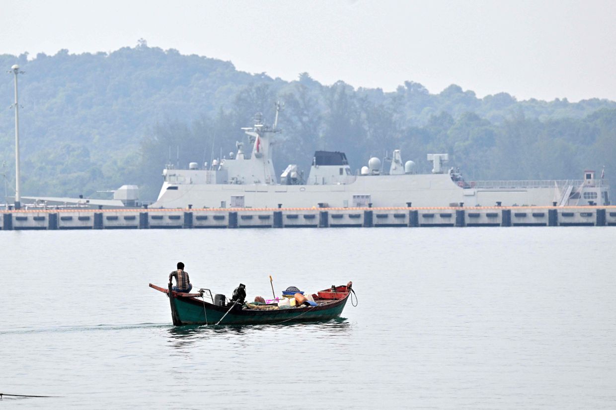 A boatman looks on from his skiff as a Chinese navy ship is seen in the background at the Ream Naval base, located in Cambodia's southern coast in Preah Sihanouk province, on January 24, 2026. The US Navy's USS Cincinnati (LCS-20) littoral combat ship, on Saturday, January 24, is also on a port call to the Cambodian naval base. -- Photo by TANG CHHIN Sothy / AFP