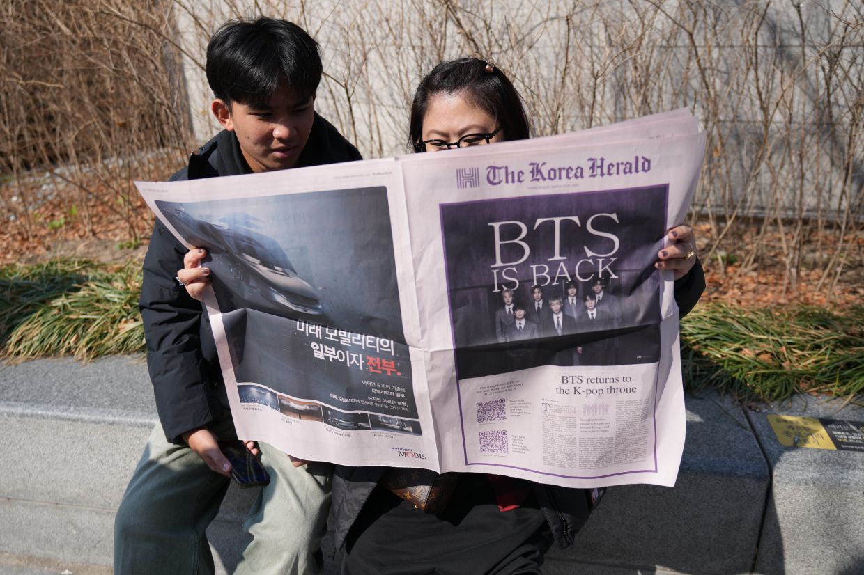 Tourists from Singapore read special edition of The Korea Herald newspaper reporting about BTS near a stage for the comeback concert of K-pop band BTS at Gwanghwamun Square in Seoul, South Korea, Saturday, March 21, 2026. (AP Photo/Lee Jin-man)