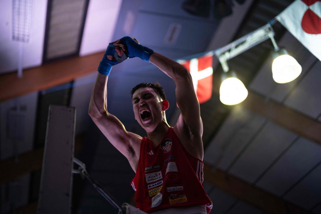 Nunu, a young Greenlandic fighter, celebrates while raising a trophy after a bout against a Danish opponent, with the Danish and Greenlandic flags in the background, during a tournament in Nuuk, Greenland, on February 28, 2026.