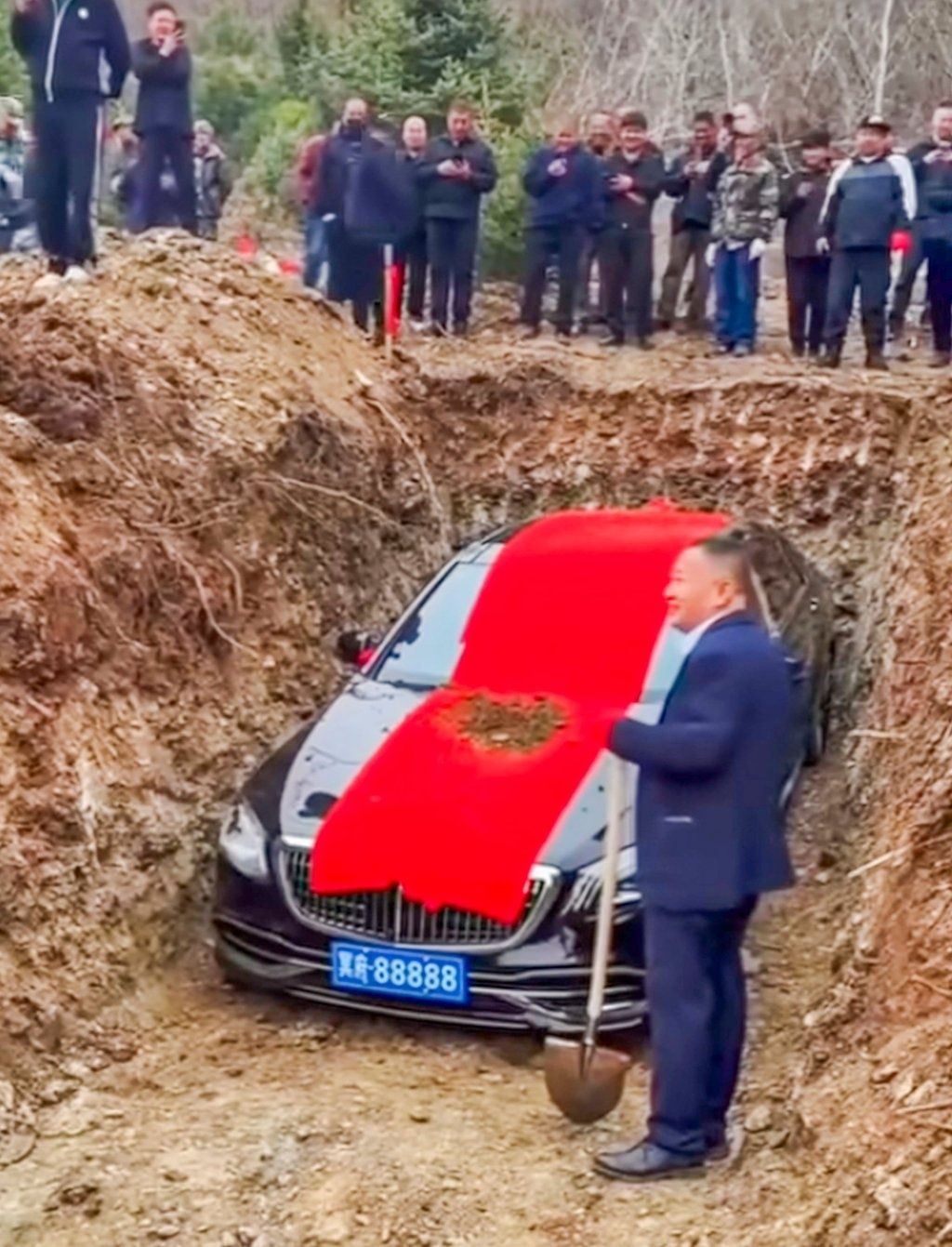 A mourner stands next to the about to be buried luxury car holding a spade. - Photo: bilibili.com