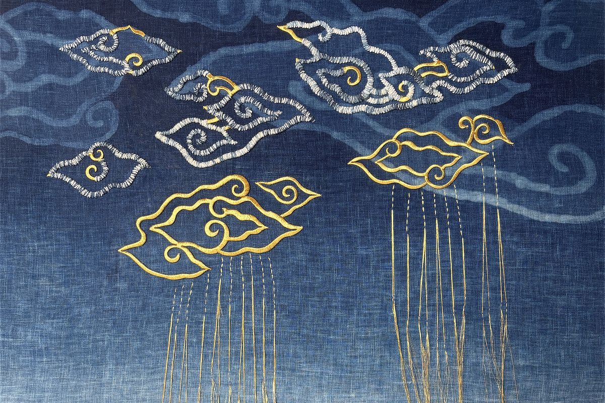 Textile artist Yann tilts her head at her work Blessing, where clouds and fine golden threads fall over an indigo batik linen backdrop.