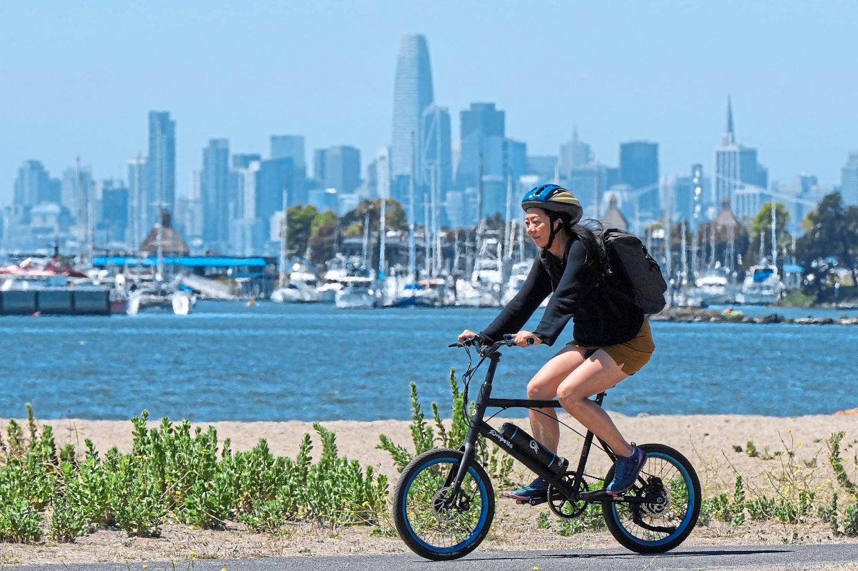 A woman rides a bicycle on a path, as the San Francisco skyline is seen in the background, in Alameda, California. 