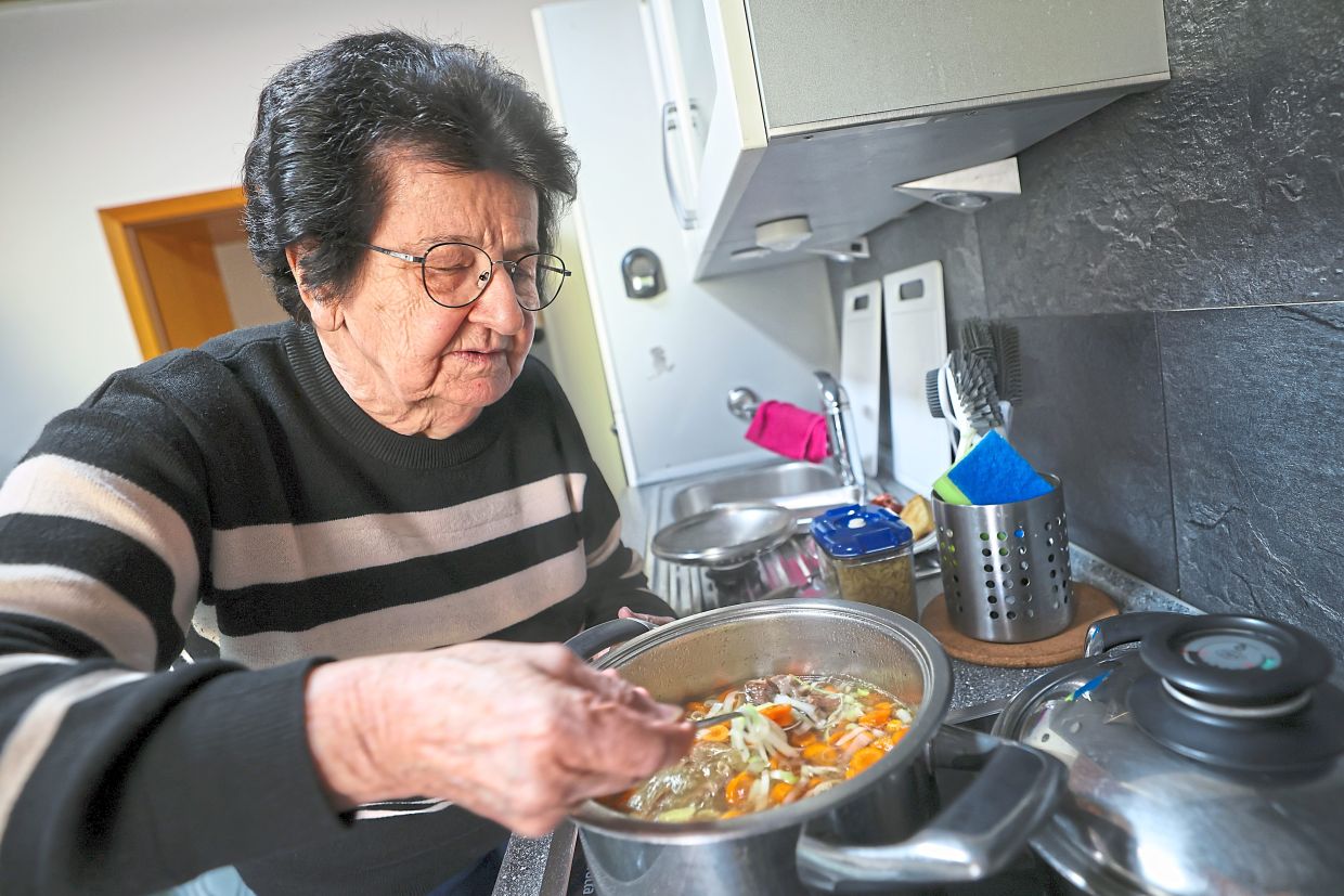 Annemarie, stirring a pot filled with vegetables and beef, does the cooking while her granddaughter films.