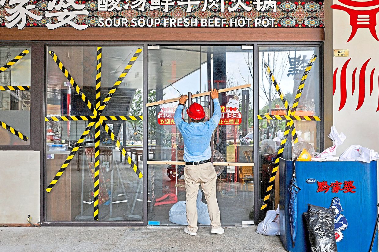 Weather woes: A man securing the door of a taped-up restaurant with wooden bars in Wenzhou, Zhejiang province, China; and (below) pedestrians wearing raincoats making their way through strong winds in Ishigaki, Japan. — Reuters