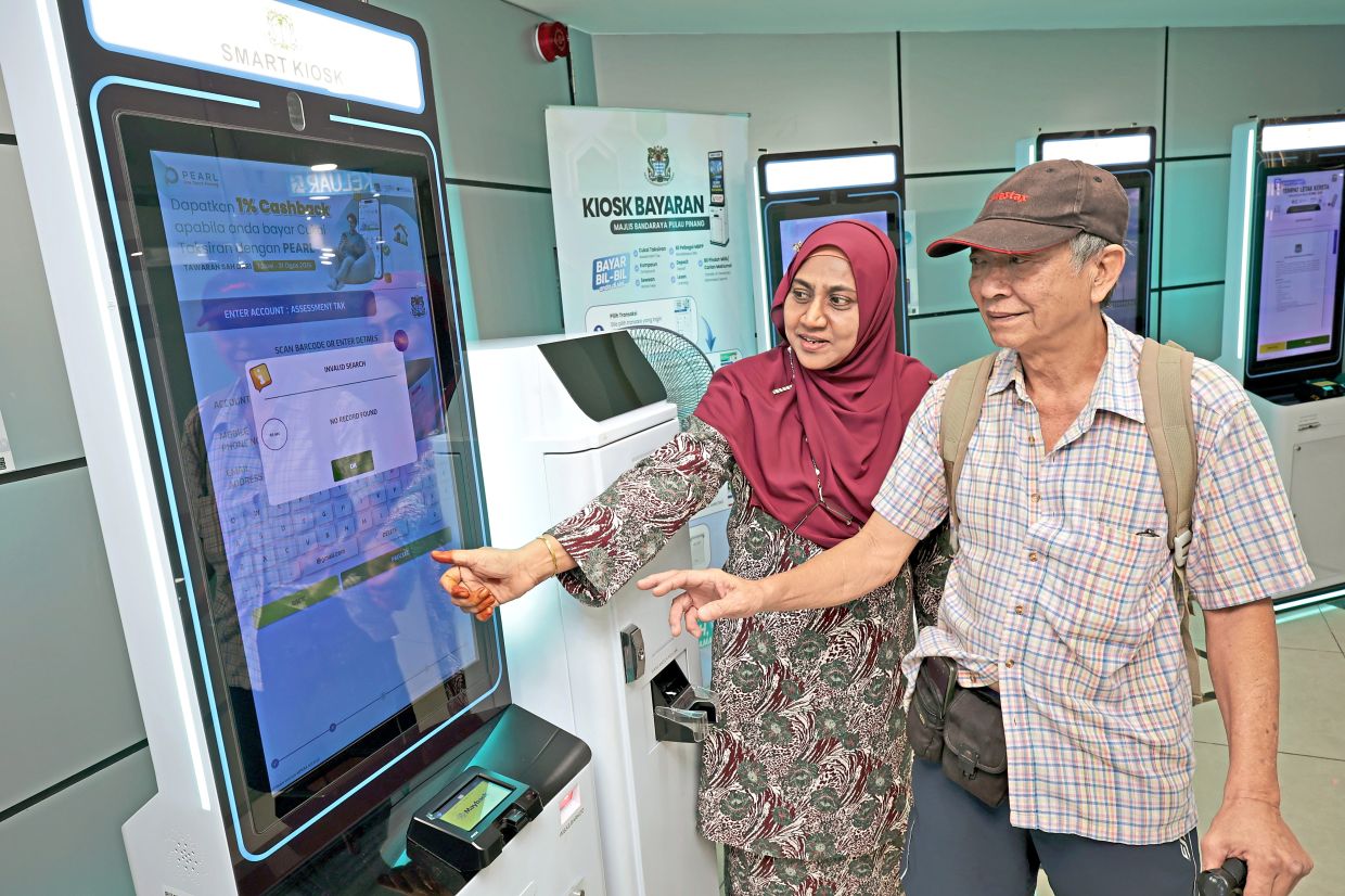 A senior citizen getting assistance from a Penang Island City Council employee on how to make a cashless transaction at a kiosk in Komtar.
