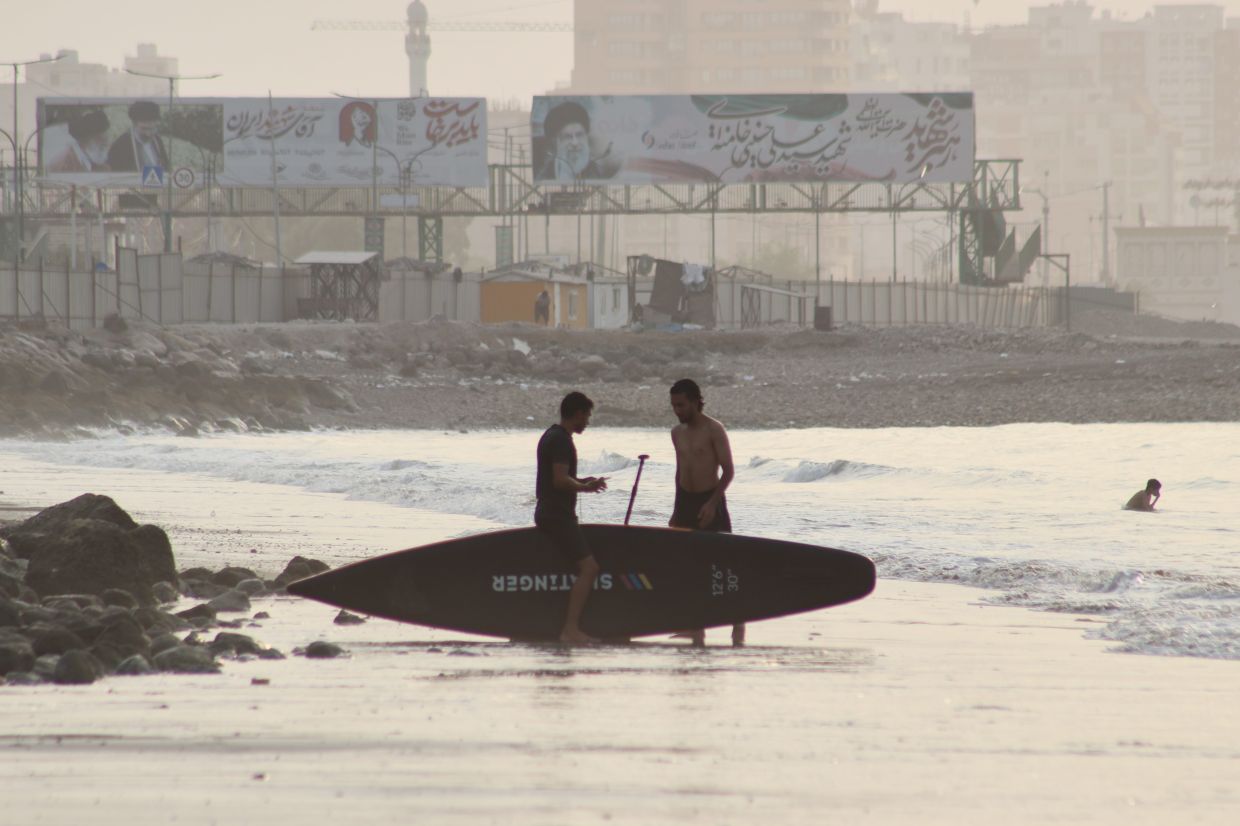 Two men stand with a surfboard along the shore in the Strait of Hormuz off Bandar Abbas, Iran, as billboards depicting Iran's late Supreme Leader Ayatollah Ali Khamenei rise in the background, Monday, July 27, 2026. (Razieh Poudat/ISNA via AP)