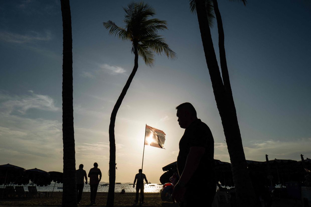 People walk past a national flag of Thailand fluttering in the wind on Pattaya Beach in Pattaya on September 1, 2026. Thailand will halve the length of visa-free stays for tourists from more than 60 countries and territories to 30 days, effective September 15, the foreign ministry said on September 1. (Photo by ANTHONY WALLACE / AFP)