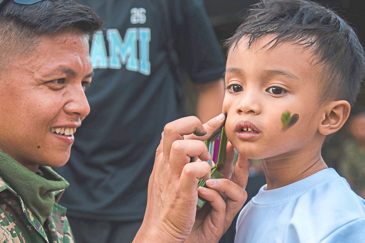 A member of the Malaysian Armed Forces paints hearts on a child’s face, adding a tender touch to the National Day festivities.