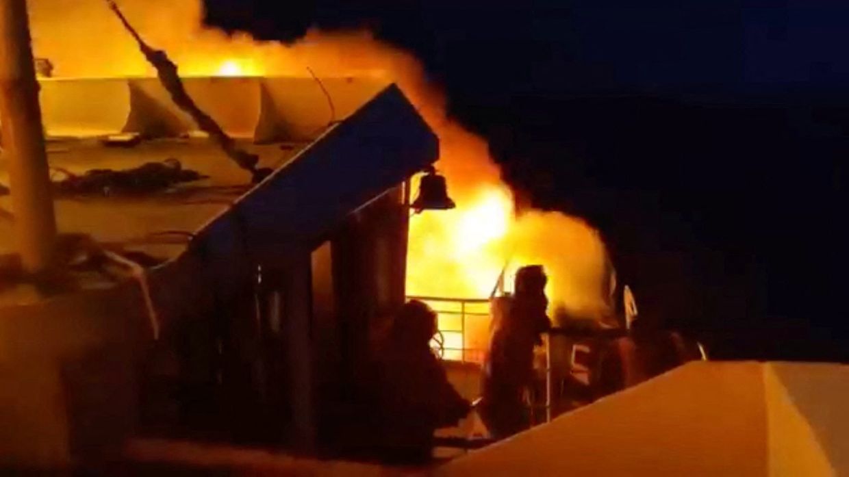 People stand aboard MV June Aster as flames and smoke rise after the fire broke out. - Reuters