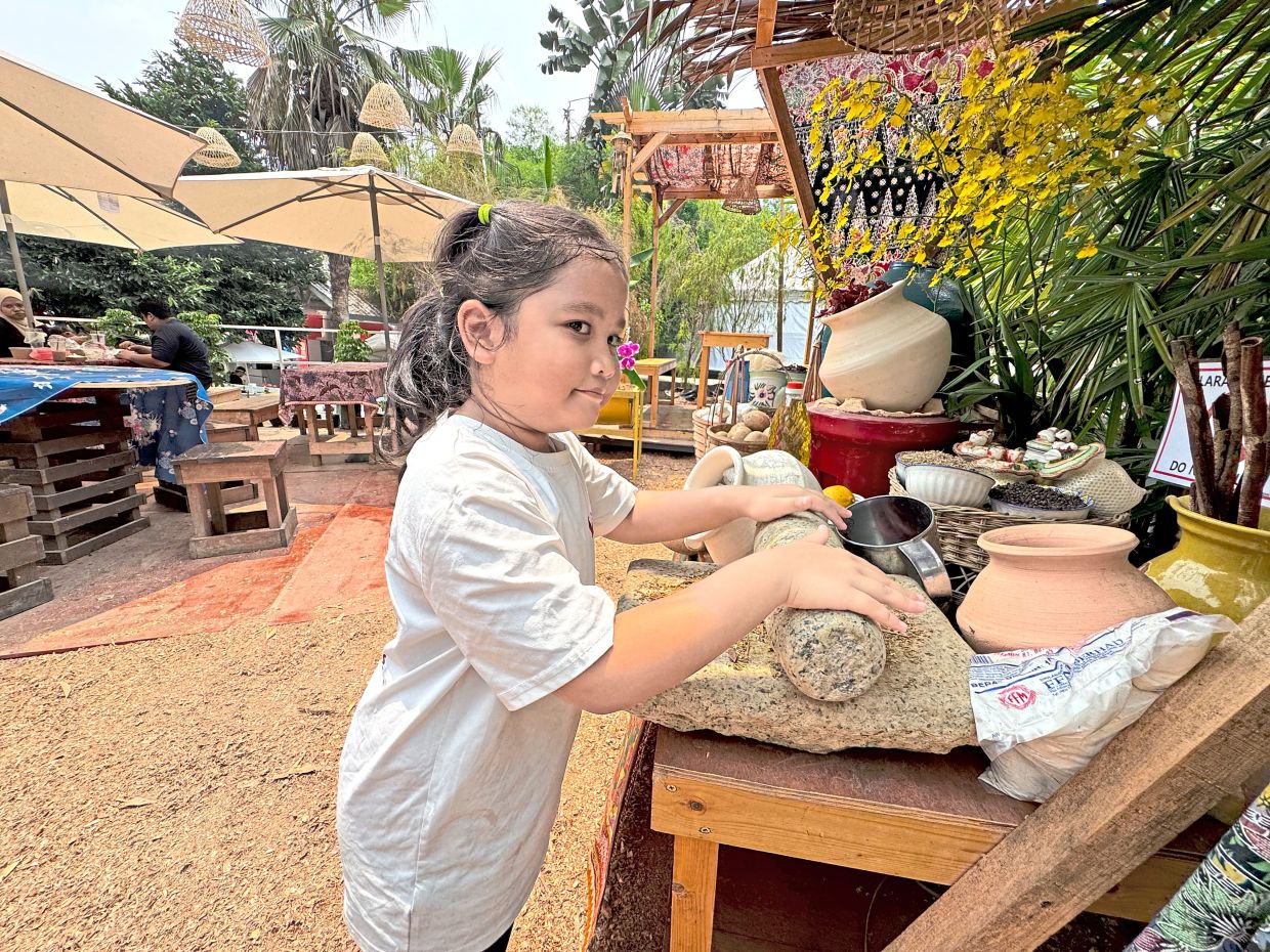 Siti Nur Sumayyah Zuhri, seven, trying a batu giling loaned by a DBKL employee for interactive display at the village kitchen.