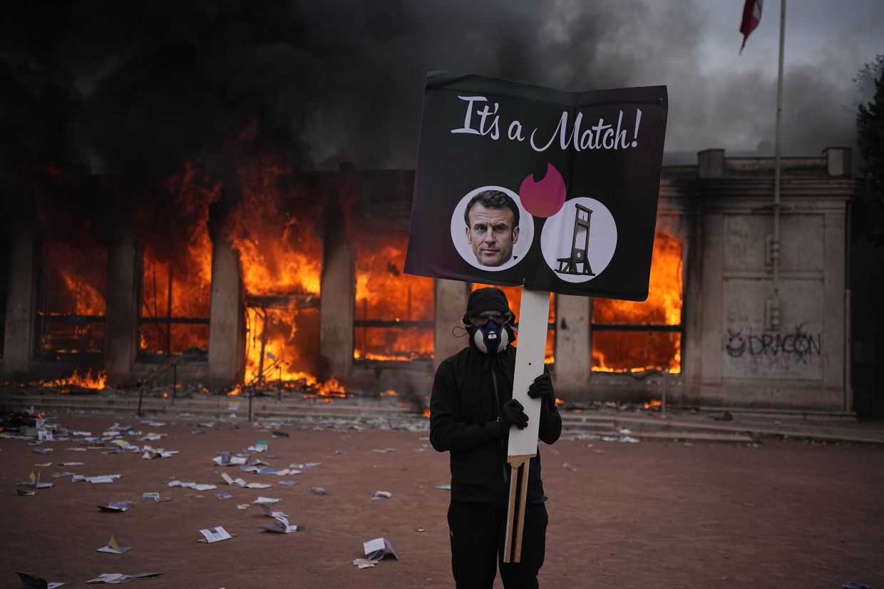A protester holds a board with a photo of French president Emmanuel Macron in front of a burning tourism office during a demonstration against rising living costs and in solidarity with students’ grievances in Lyon, France, Saturday, Oct. 17, 2026. (AP Photo/Laurent Cipriani)